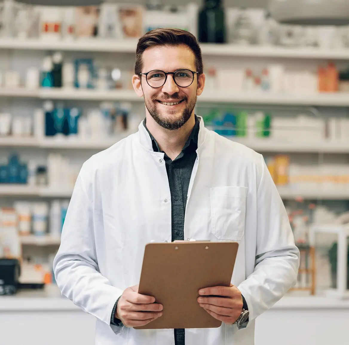 A pharmacist with glasses, wearing a white coat, holds a clipboard while standing in a pharmacy setting with shelves of products in the background.