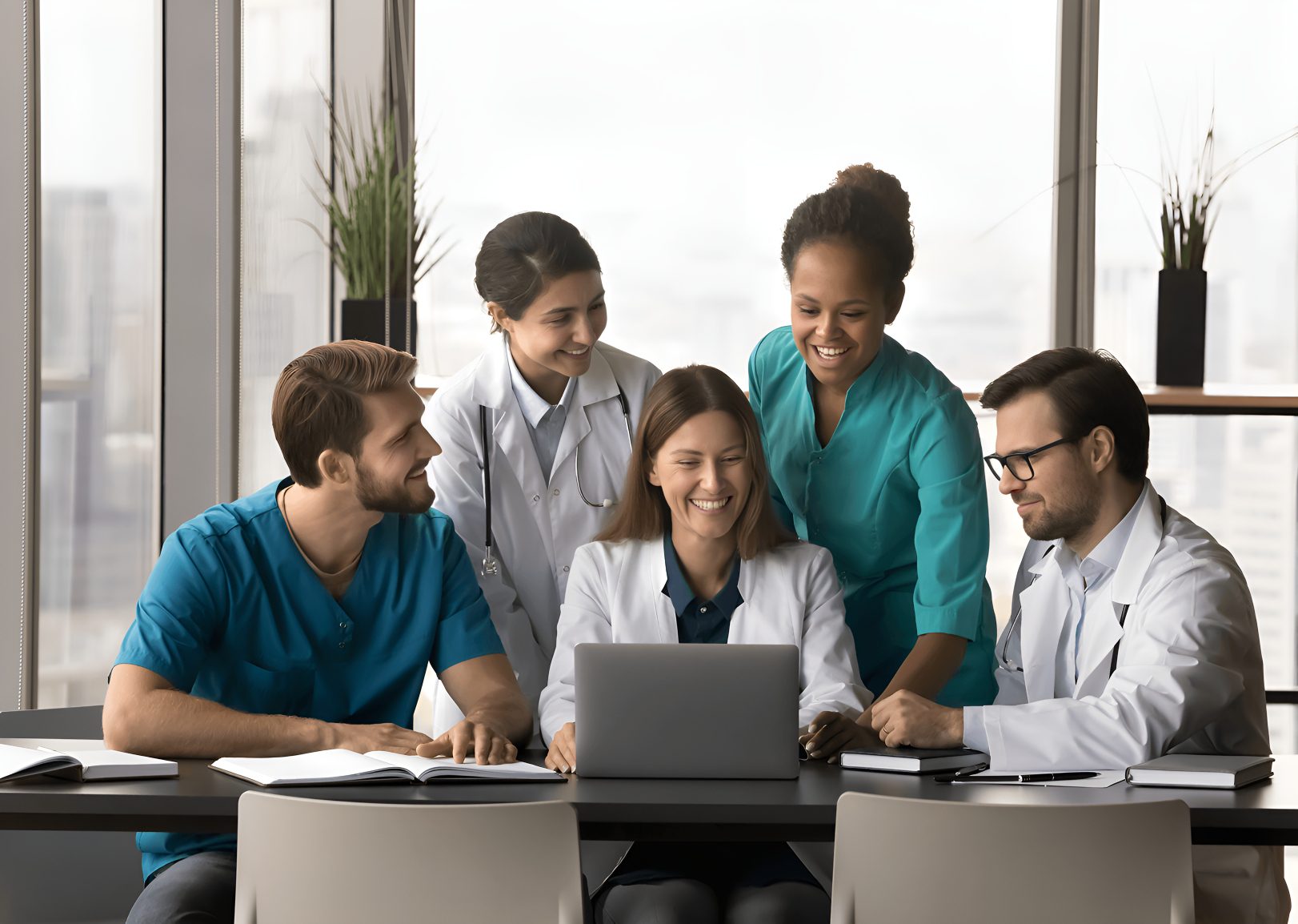 A group of healthcare professionals, wearing lab coats and scrubs, gather around a table looking at a laptop, smiling and engaged in discussion.