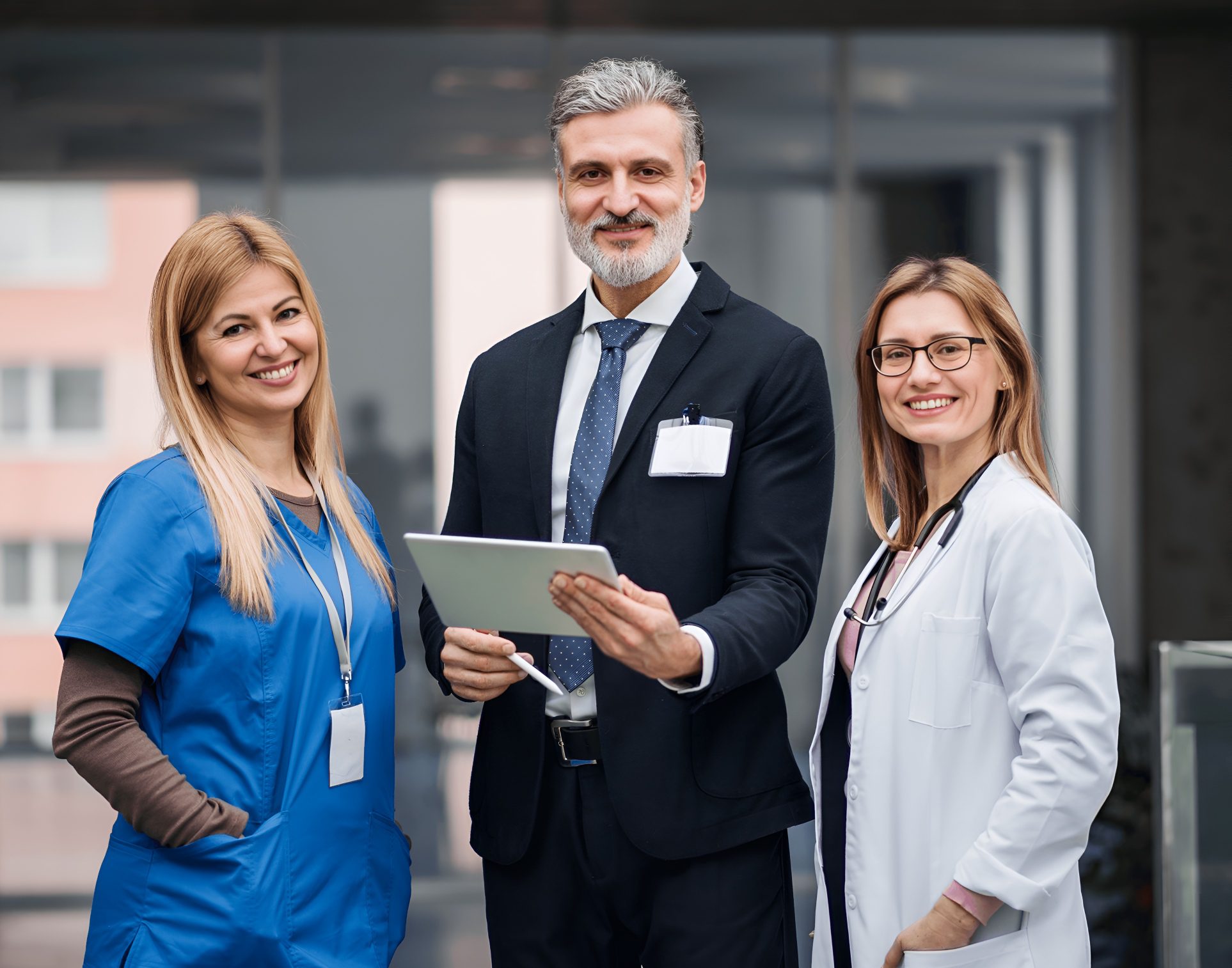 Three professionals smiling, two in medical attire and one in a suit, hold a tablet indoors.